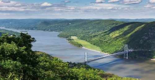 A river winding through a lush, green valley with a suspension bridge spanning it. The landscape, reminiscent of the Highlands, is covered in greenery under a partly cloudy sky.