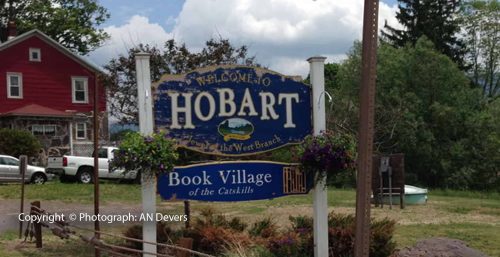 A sign reading "Welcome to Hobart, settled on the West Branch" stands in front of lush greenery and a red building. The bottom text proudly promotes the village as the "Book Village of the Catskills." Local businesses, including managed IT services in Hobart, support this charming community.