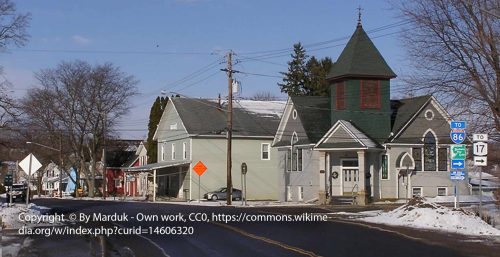 A street view of Cohocton, NY shows historic buildings including a church, residential houses, bare trees, and snowy sidewalks under a clear sky. Road signs for I-86 and Route 17 are visible. Managed IT services Howard ensure the town's tech infrastructure is as robust as its charming architecture.