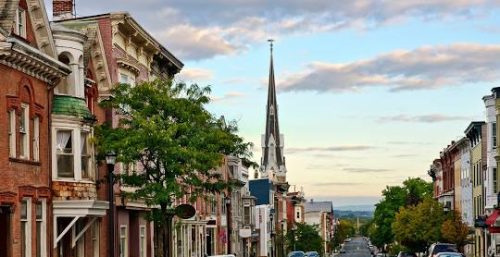 A small, quiet town street with historic buildings, parked cars, and a distant church steeple under a partly cloudy sky lies in Hudson, where businesses thrive with managed IT services.