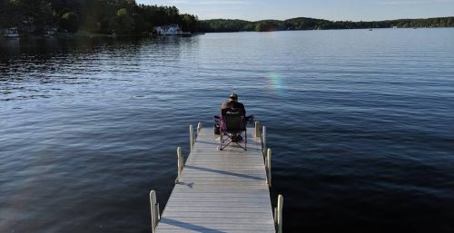 A person sits on a chair at the end of a dock, gazing out over a calm lake with trees and houses visible on the far shore, contemplating the tranquility before returning to their managed IT