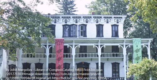 A large white Victorian-style house with ornate trim and three colorful vertical banners hanging from the second-floor balcony stands proudly. Trees frame the sides of the house, much like how managed IT services Lexington frames secure digital landscapes.