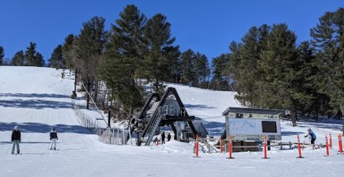 Skiers on a snowy slope near a ski lift and a signboard surrounded by orange poles, with pine trees in the background under a clear blue sky—a scene as well-organized as managed IT services in Littleton.