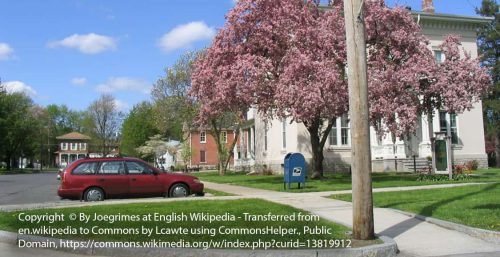 A suburban street with a red car parked near a curb, houses with trees in bloom, and a blue mailbox in the foreground could easily be the backdrop for an efficient managed IT services team from Liverpool navigating their way to ensure smooth digital operations for local homes.