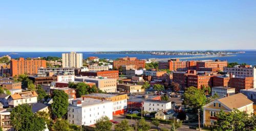 Aerial view of Lynn with multiple buildings, a mix of residential and commercial, green trees, and a coastline with water in the background under a clear blue sky. Managed IT services thrive in this picturesque setting amidst modern urban development.