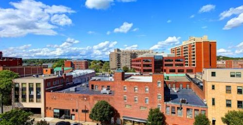 A cityscape view featuring a mix of low-rise brick buildings and a taller, modern apartment block under a clear blue sky with some clouds. Trees line the streets below, hinting at the harmony between technology and nature in Malden, where managed IT services flourish alongside urban development.