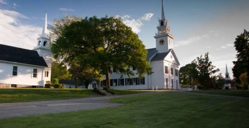 A large white church with a clock tower and steeple stands in a grassy area with trees, adjacent to another smaller white building with a similar steeple, reminiscent of the charm you'd find in Milton. The sky is partly cloudy.