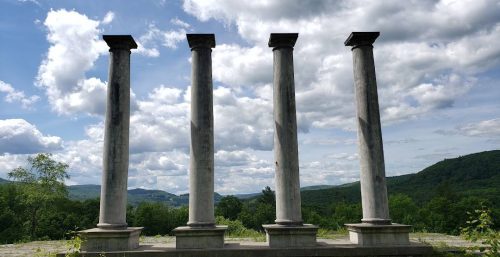 Four tall stone columns stand in a row against a backdrop of a cloudy sky and the green, hilly landscape of Monterey.