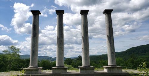 Four tall stone columns stand in a row against a backdrop of a cloudy sky and the green, hilly landscape of Monterey. Four tall stone columns stand in a row against a backdrop of a cloudy sky and the green, hilly landscape of Monterey.
