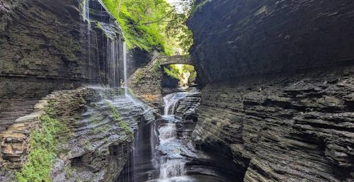 A narrow gorge with multiple small waterfalls cascading down layered rock formations. A stone bridge arches over the gorge, surrounded by lush greenery, reminiscent of the tranquility one might seek after a day of managed IT services in Odessa.