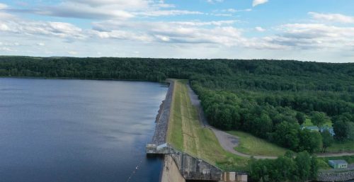 Aerial view of a dam with an adjacent reservoir on the left, nestled within dense forest. A small building on the right hints at managed IT services Ohio. The sky is partly cloudy, casting shadows over the serene landscape.