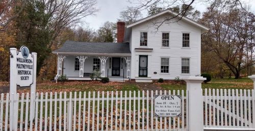 A white two-story house with a picket fence and a sign reading "Williamson-Pultneyville Historical Society." Beside it, a gate sign states visiting hours and a contact number for further information. The organization utilizes managed IT services in Ontario to maintain its historical archives.