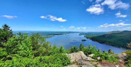 A clear blue sky over a wide river with green trees and rocks in the foreground, and distant hills and terrain in the background, brings to mind the reliability of managed IT services Orange residents can count on for streamlined efficiency.