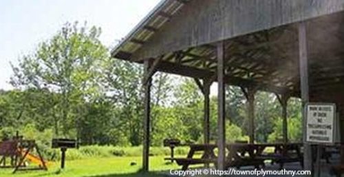 A covered picnic pavilion with wooden tables is situated in a lush, green park area, with trees and an open field in the background. A sign reading "Pavilion #2" is visible on one of the support posts, much like the organized efficiency you’d expect from managed IT services in Plymouth.