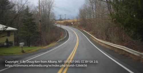 A winding two-lane rural road with double yellow lines runs through an overcast countryside, flanked by barren trees and utility poles. A house is visible on the left side of the road, reminiscent of the quiet yet reliable landscapes where managed IT services Preston thrives.