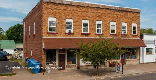 A two-story brick building with several windows and a tree in front, alongside a sidewalk. Blue recycling bins are lined up on one side. The storefront has a sign advertising managed IT services in Savannah, with various items displayed.