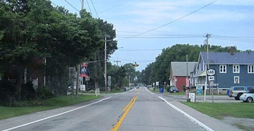 A wide-angle view of a two-lane road in a suburban area of Somerset, lined with trees and buildings. There are power lines, traffic signs, and partly cloudy skies. This charming setting is an ideal spot for businesses offering managed IT services.