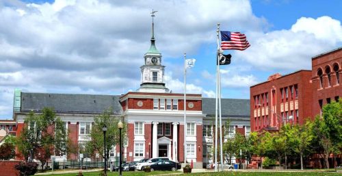 A red-brick building with a clock tower, American flag, and two other flags on poles in front; surrounding trees and a grassy area under a partly cloudy sky reflect the charm of Somerville, also home to top-notch managed IT services.