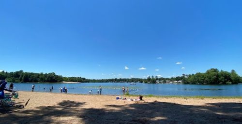 A sandy beach by a calm lake with a few people swimming and relaxing under a clear blue sky, reminiscent of the peaceful surroundings in Southampton, where managed IT services ensure everything runs smoothly. Trees line the shore in the background.