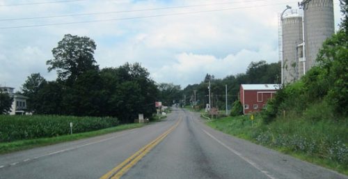 A rural road with double yellow lines extends into the distance, flanked by trees, a cornfield on the left, and a red barn with silos on the right under a partly cloudy sky—a perfect drive for anyone in Springfield offering managed IT services to clear their mind.