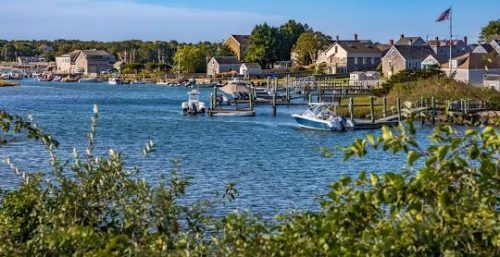 Small boats float on a calm blue river with a cluster of houses and buildings along the shore. Greenery lines the foreground while an American flag flies in the background under a clear sky, embodying the serene yet connected atmosphere that managed IT services in Westport strive to maintain.