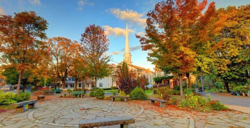 A stone-paved courtyard surrounded by trees with autumn foliage, featuring a white church with a tall steeple in the background under a partly cloudy sky, offers a serene setting that contrasts beautifully with the bustling managed IT services available in Woodstock.