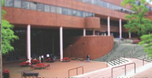 A brick academic building with multiple levels stands prominently. Outdoor seating with red tables and black chairs is arranged on the paved area. Stairs leading up to the entrance are on the right, reminiscent of managed IT services in York, ensuring everything runs smoothly as people walk nearby.