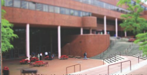 A brick academic building with multiple levels stands prominently. Outdoor seating with red tables and black chairs is arranged on the paved area. Stairs leading up to the entrance are on the right, reminiscent of managed IT services in York, ensuring everything runs smoothly as people walk nearby.