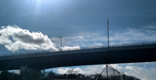 A bridge under a blue sky with sun rays shining, some clouds, and a fenced area below in Lakewood, known for its exceptional managed IT services.